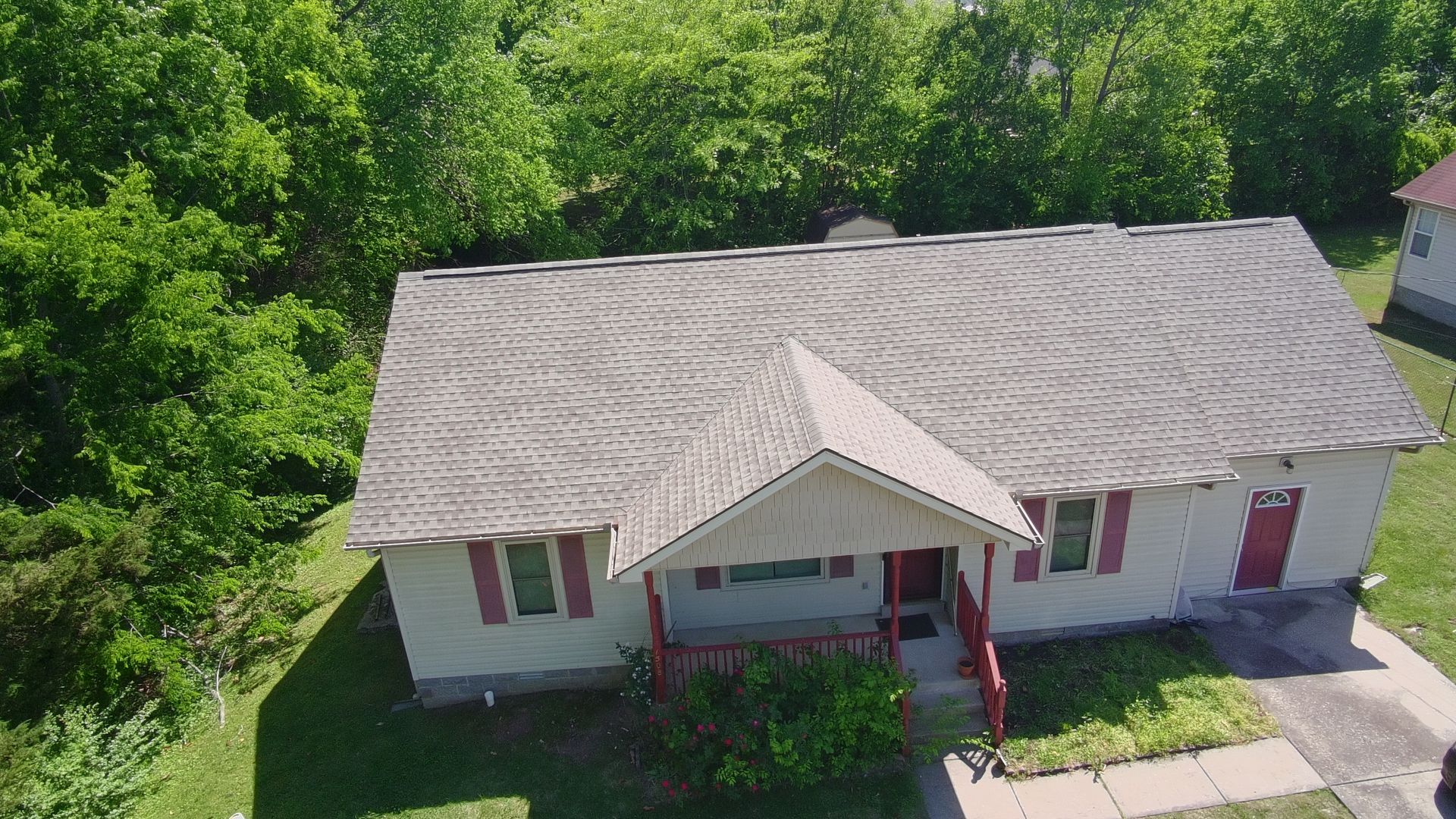 Aerial view of a single-story white house with a gray roof, red shutters, and a front porch surrounded by trees.