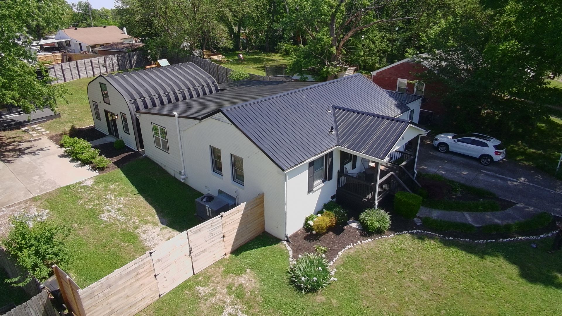 Aerial view of a white house with a dark roof, surrounded by trees, lawn, and a driveway with parked cars.