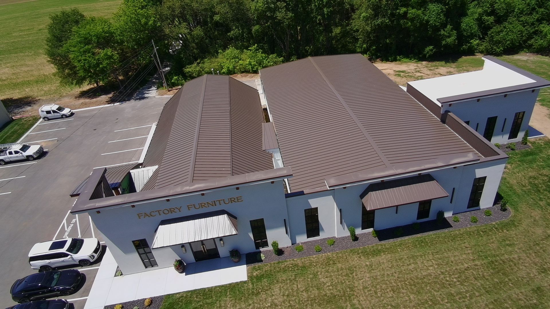 Aerial view of a large white building with a wavy brown roof, parked cars, and surrounding trees