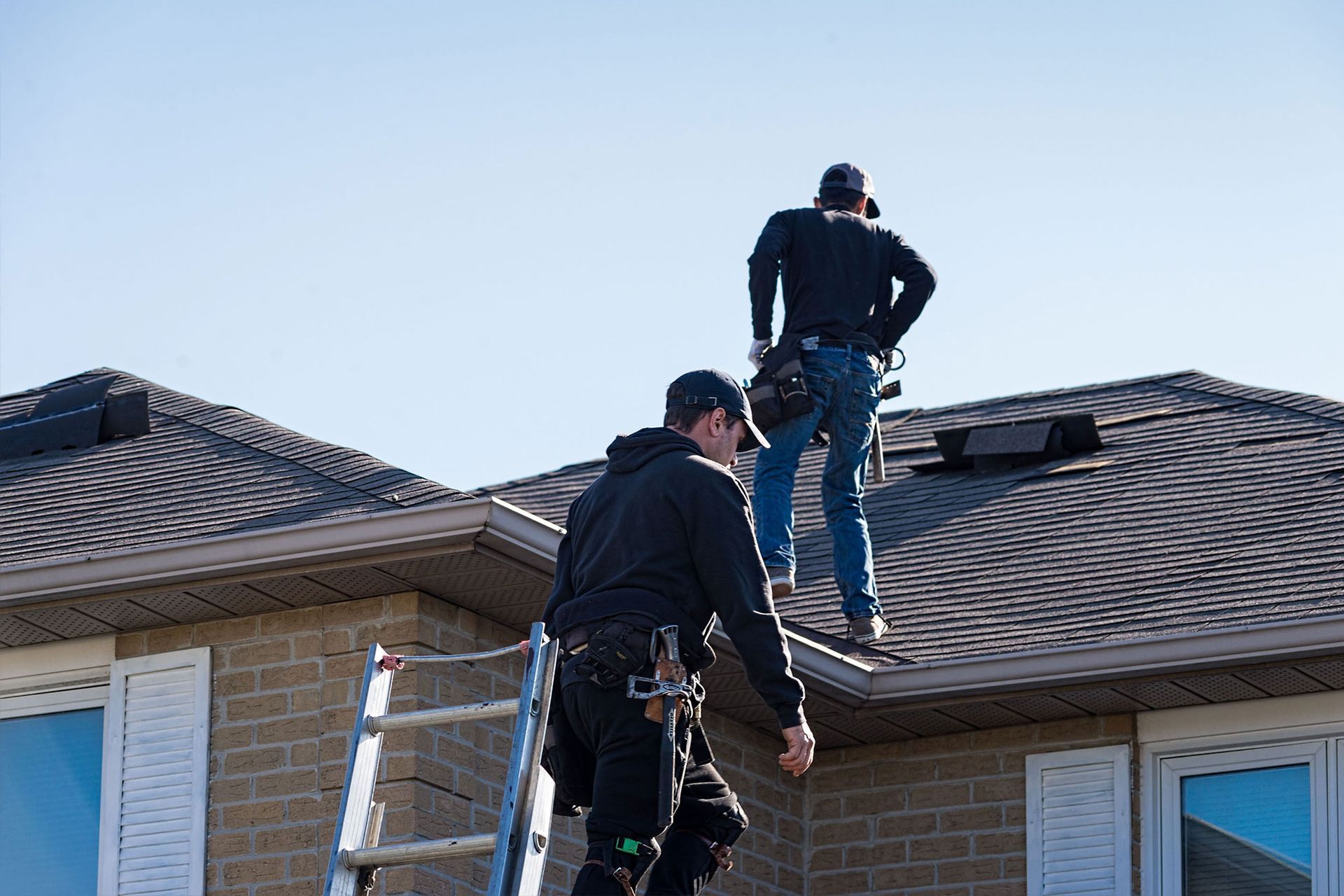 Two roofers working on a shingled house roof, one standing on a ladder below the other.