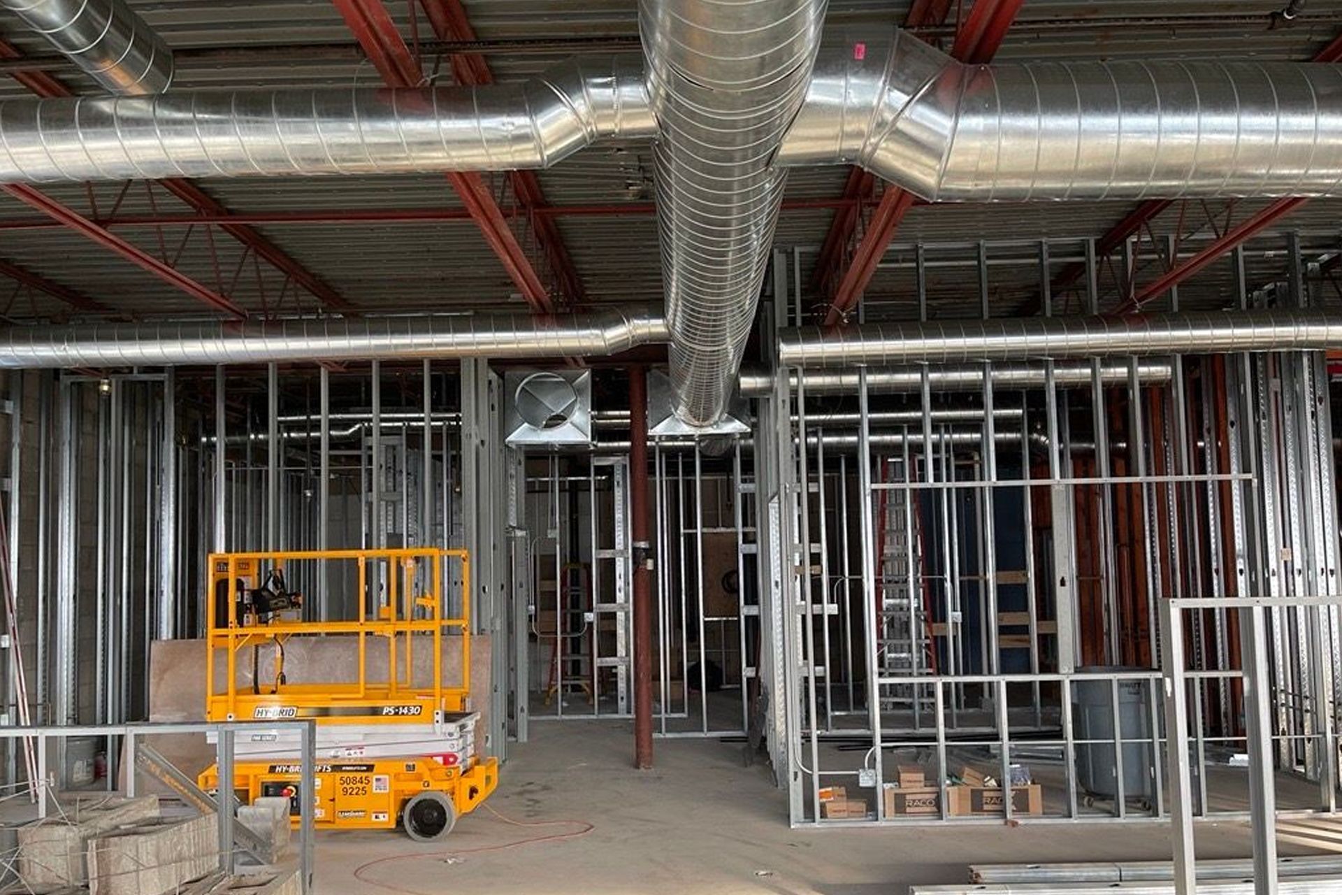 Interior construction site with metal framing, HVAC ductwork, and a yellow scissor lift.