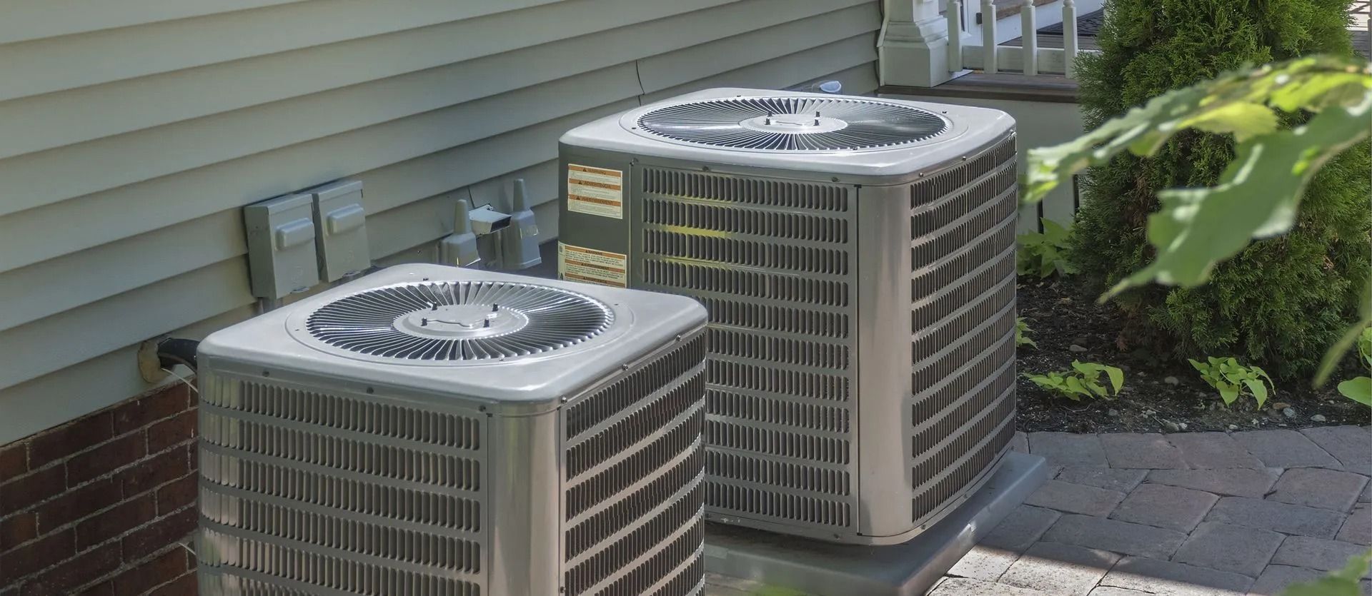 Two air conditioning units sit on a patio next to a house with brick and siding.