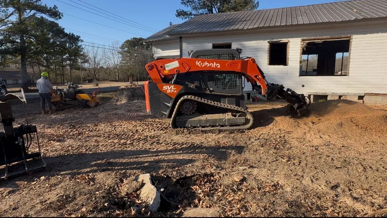 An orange Kubota track loader operates near a white building with missing windows on a dirt lot.