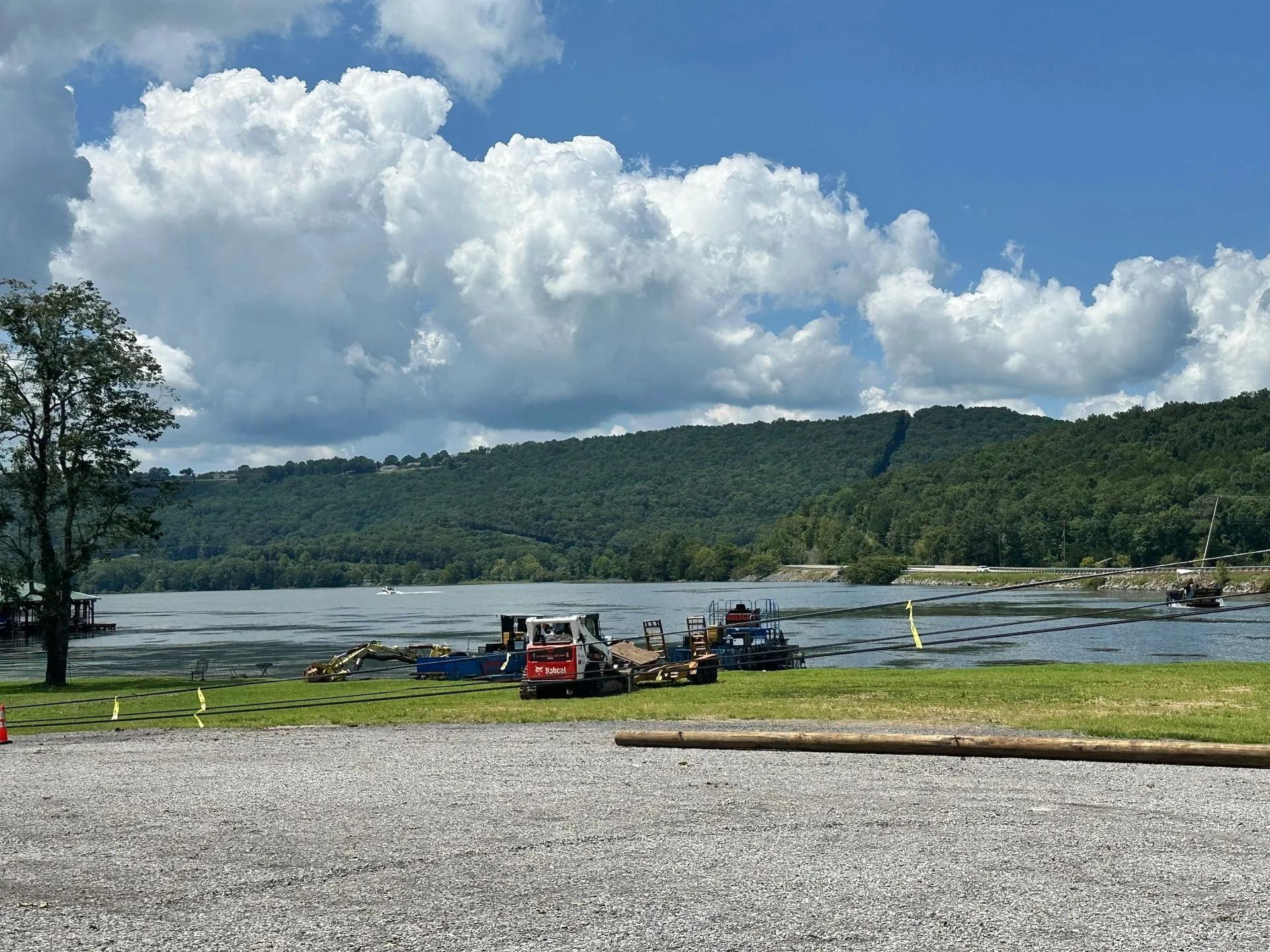A red skid steer loader parked on a grassy lakeshore with a forested mountain and cloudy blue sky in the background.