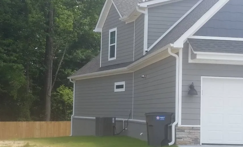 Gray house side with white garage door, black trash bin, and wooded backyard fence.