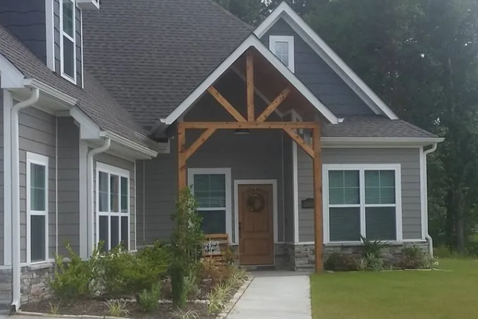 Covered front entrance of a gray suburban house with wood trusses and a concrete walkway.