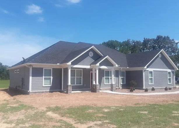 Gray single-story house with dark roof and white trim on a dirt lot under a blue sky
