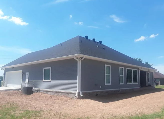 Gray single-story house with a dark roof and white trim on a dirt lot under a blue sky.