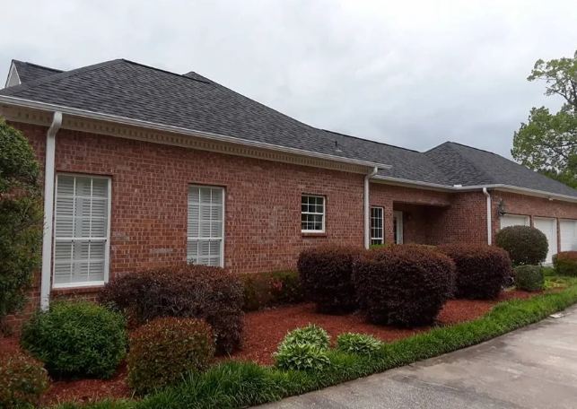 Brick house exterior with dark roof, trimmed shrubs, and a driveway on a cloudy day