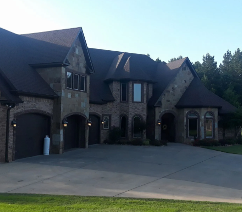 Large stone suburban house with arched garage doors and a wide concrete driveway under a blue sky