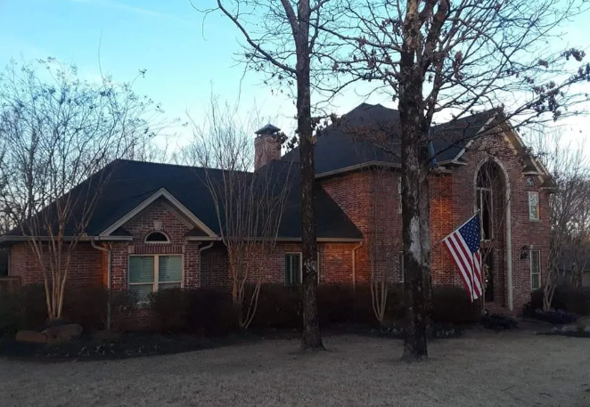 Large brick suburban house with an American flag, leafless trees, and a gravel driveway at dusk