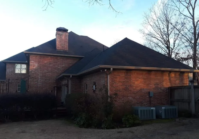 Brick house with a dark roof, chimney, and bare trees in the yard at dusk
