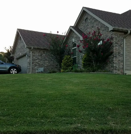 Stone house with a steep roof and flowering bushes behind a green lawn, with a car at left.
