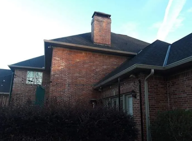 Brick suburban house with dark roof and chimney, partly obscured by bushes