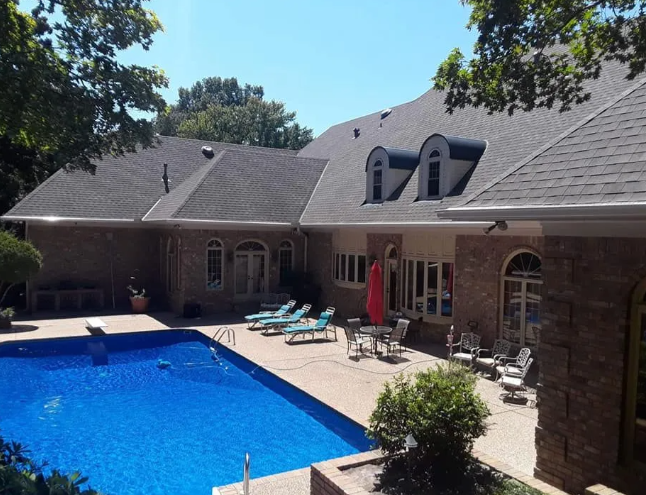 Backyard patio with a blue swimming pool, lounge chairs, and a brick house with dormer windows