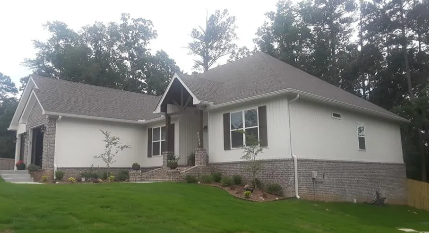 Single-story suburban house with gray siding, stone foundation, and a front porch on a green lawn.