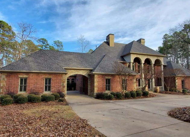 Large brick house with a gabled roof, arched entryway, and driveway surrounded by trees