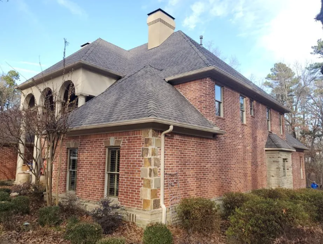 Brick house with stone accents and dark shingled roof surrounded by leafless shrubs and trees