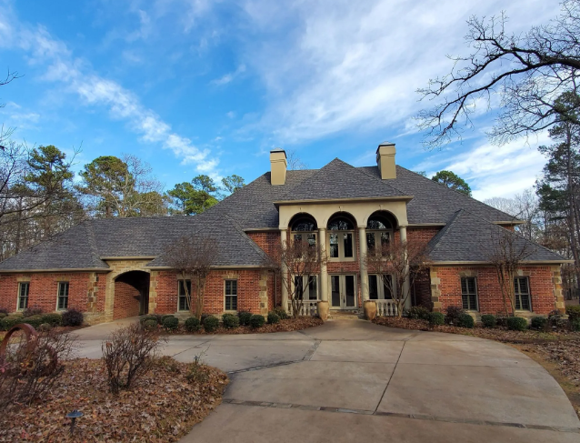 Large brick suburban house with arched front porch, two chimneys, and a curved driveway under a blue sky