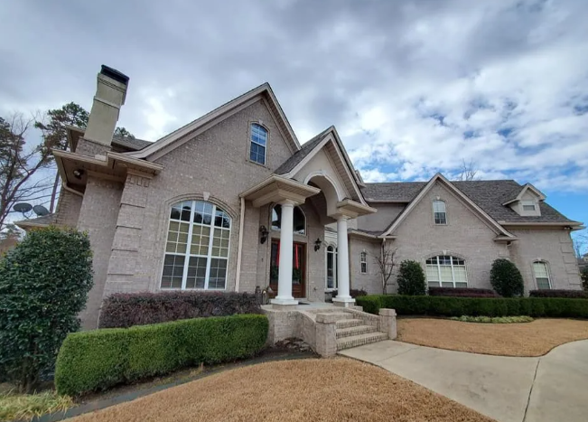 Large brick suburban house with a covered front porch, driveway, and trimmed shrubs under cloudy skies