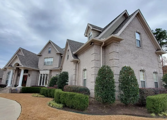 Large beige suburban house with manicured shrubs and a covered front porch under a cloudy sky