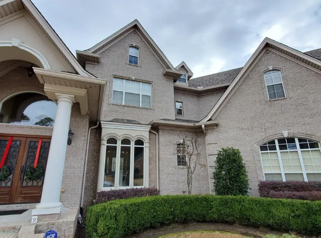 Large stone suburban house with arched windows, a front porch, and trimmed hedges under an overcast sky