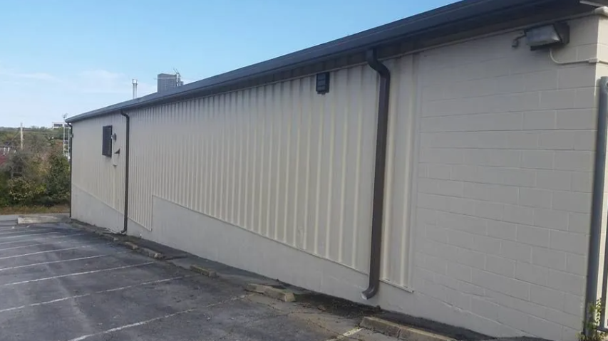 Side view of a beige building with vertical siding and a dark roof beside an empty paved lot.