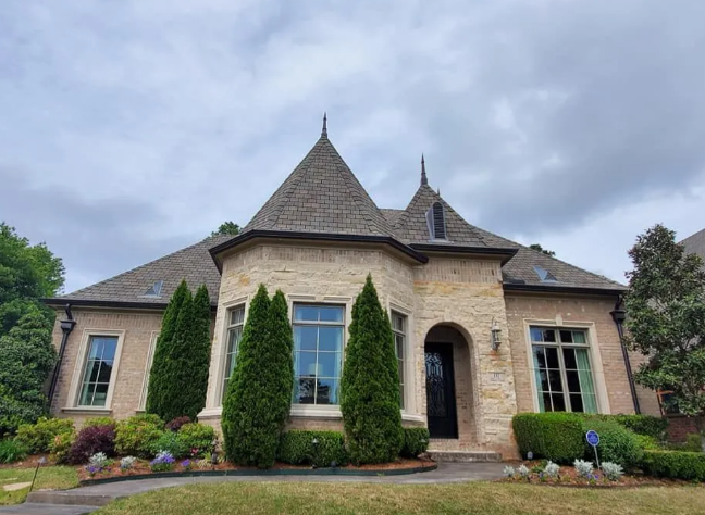 Stone suburban house with a turret, tall windows, and manicured landscaping under a cloudy sky