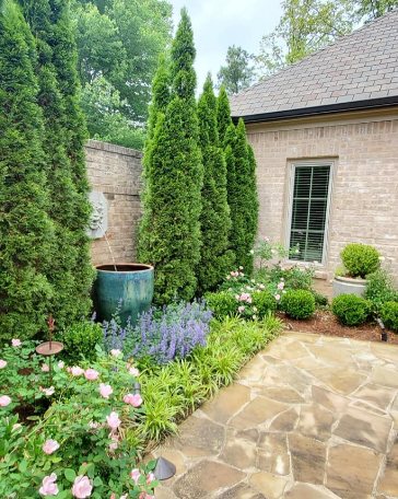 Stone patio beside a brick house, with lush garden beds, evergreen shrubs, and a blue planter fountain.