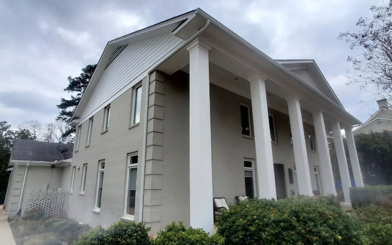 Large white neoclassical house with tall columns and shrubs under a cloudy sky