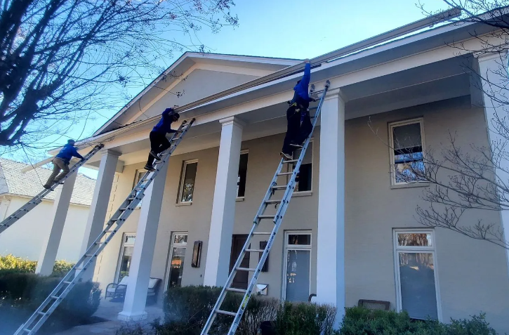Workers on ladders painting a white columned building exterior under a clear blue sky