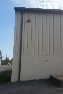 Side of a beige building with vertical siding and a small wall light, beside a paved area.