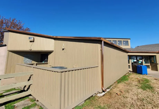Beige apartment building exterior with sloped roofs, a fenced patio, and a blue recycling bin under a clear sky