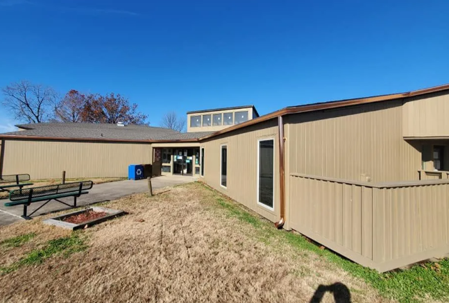 Tan building walkway leading to a doorway, with picnic tables and dry grass under a clear blue sky.