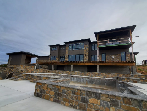 Modern stone-and-wood house with a wide deck and terraced stone retaining walls under a cloudy sky