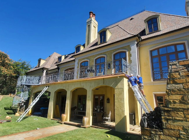 Large yellow house with balcony, stone chimneys, and blue-trimmed windows on a sunny day