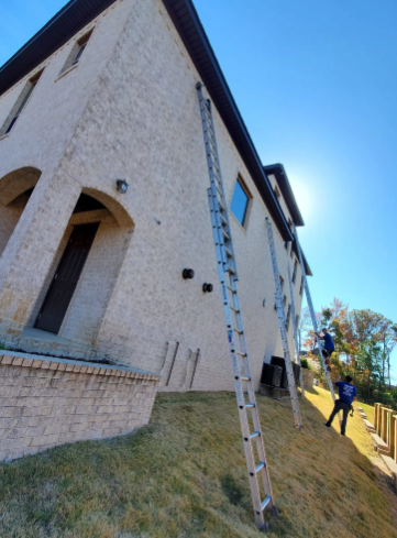 Tall beige house with a ladder leaning against it and a person standing nearby on a grassy slope.