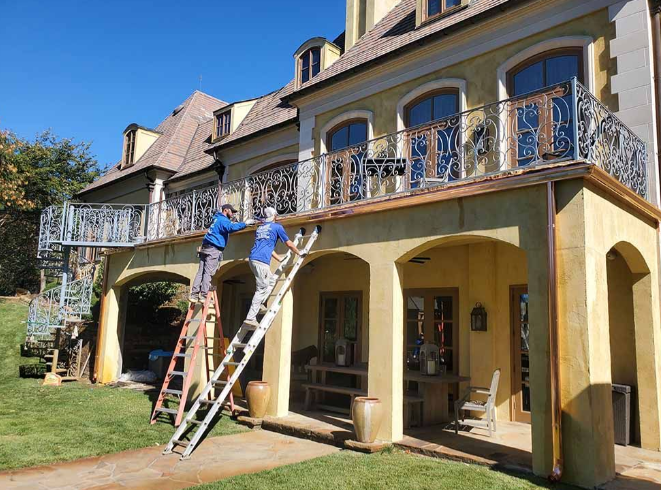 Two workers on ladders painting a house’s upper balcony and columns outdoors.