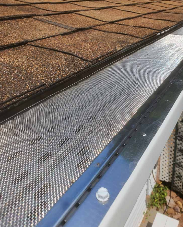 Gutter guard beneath brown shingle roof, with a blue edge and some debris below