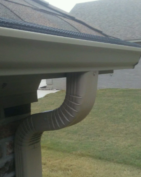 Rain gutter downspout curving from a roof eave on a house, with a grassy yard in the background