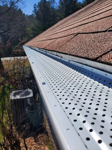 Roof edge with white perforated gutter guard under brown shingles, beside trees and a vent on a sunny day