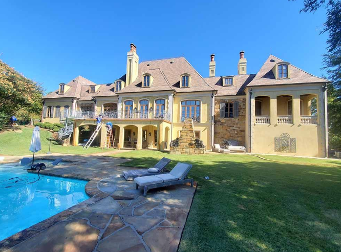 Luxury house with stone patio, blue pool, and sunny backyard lawn under a clear sky
