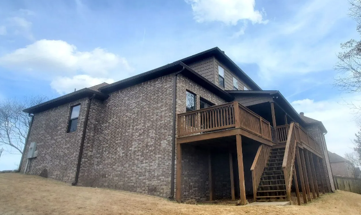 Two-story brick house with wooden deck and stairs on a grassy slope under a cloudy sky