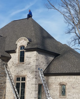 Worker on a steep gray-shingled roof beside ladders on a stone house under a blue sky.