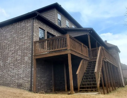 Wood-sided house with a covered deck, stairs, and support posts on a grassy slope under a cloudy sky