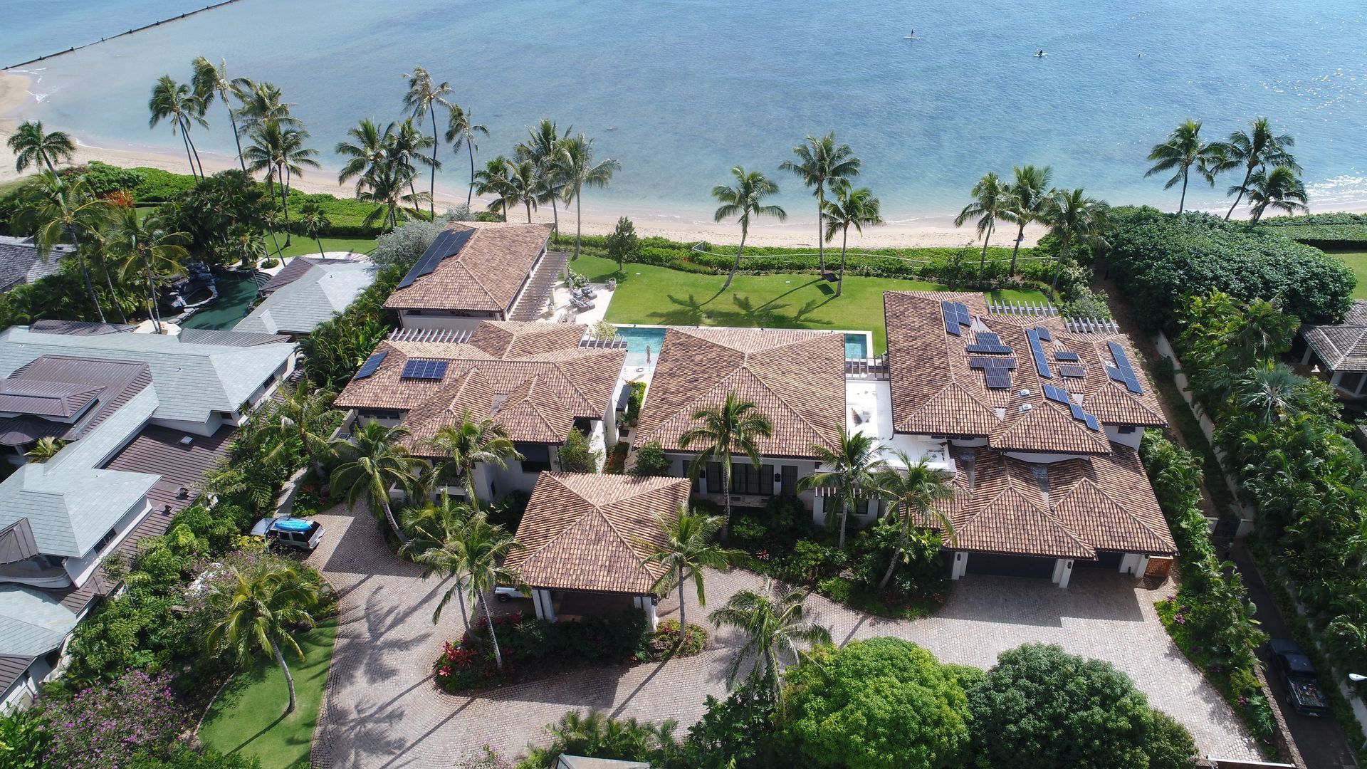 Aerial view of beachfront homes with red tile roofs, palm trees, and blue water.