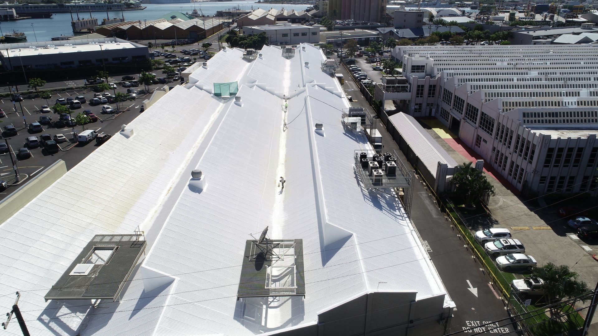 Aerial view of a white commercial roof with equipment and workers, next to a street with parked cars.