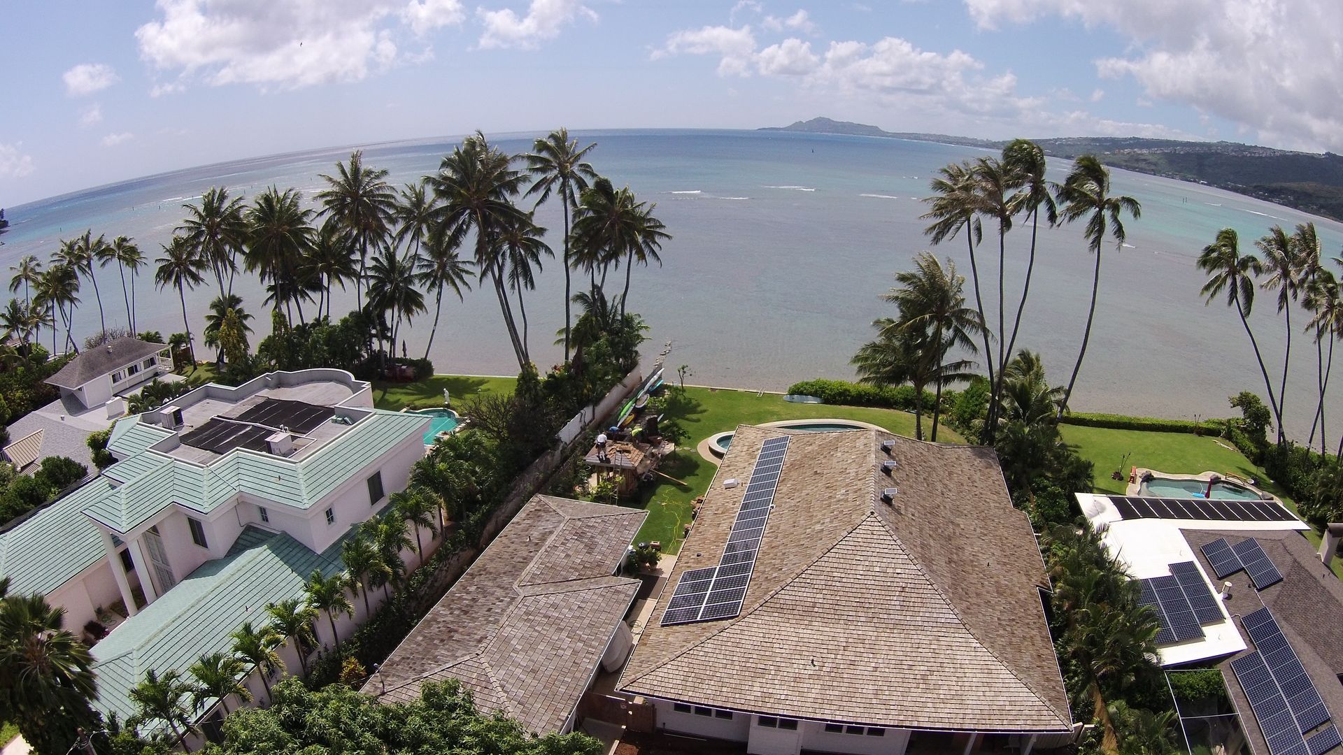 Aerial view of beach houses with solar panels, palm trees, and ocean under a blue sky.