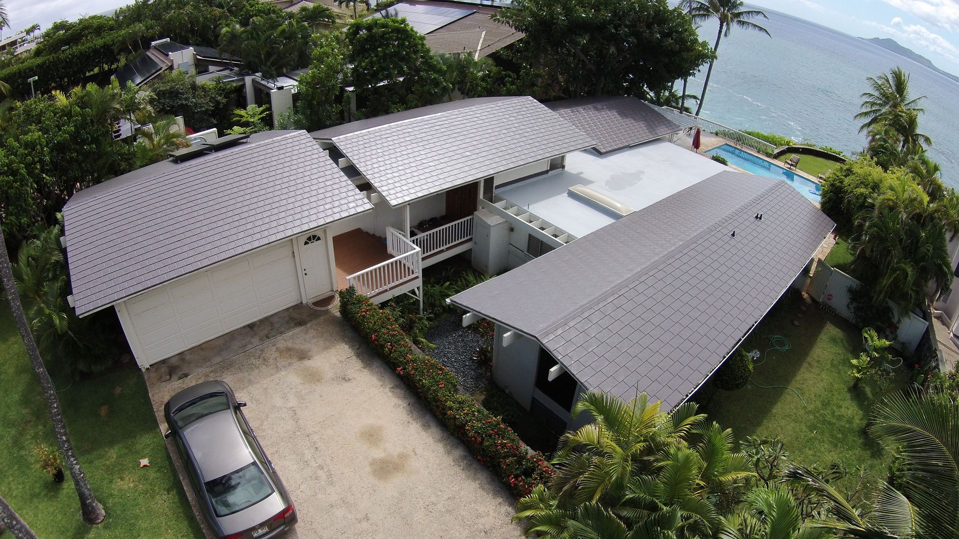 Aerial view of a white house with a gray roof near the ocean. A car sits in the driveway, surrounded by greenery.