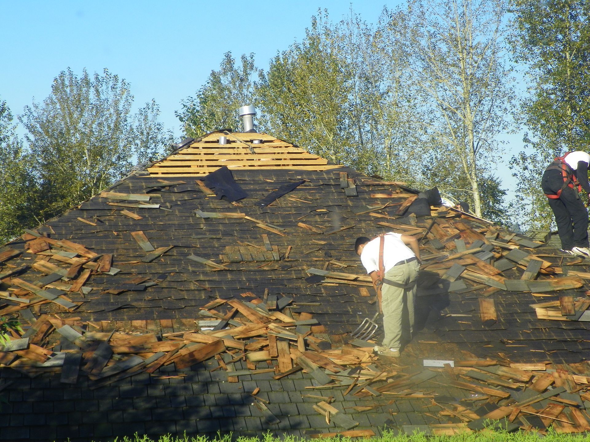 Workers removing shingles from a roof.  Debris covers the roof, with trees in the background under a blue sky.